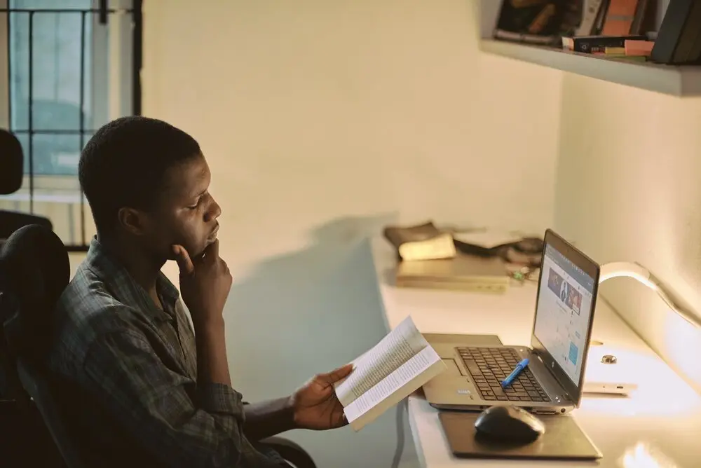 young man pondering a book as he works on a a laptop computer