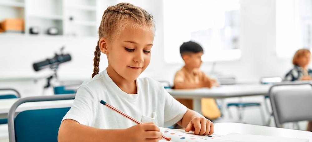 Young girl sitting at her desk, writing on a piece of paper in her classroom