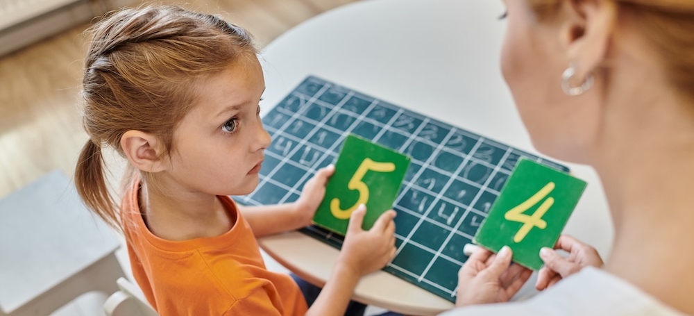 Young girl and teacher holding numbers near chalkboard, learning through play, counting, top view