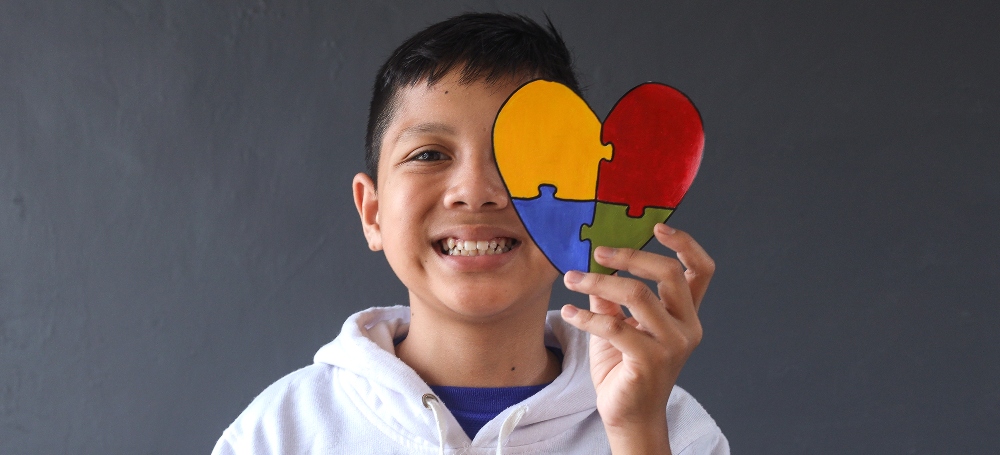 Young boy holding a heart colored in with puzzle pieces in front of his eye, smiling