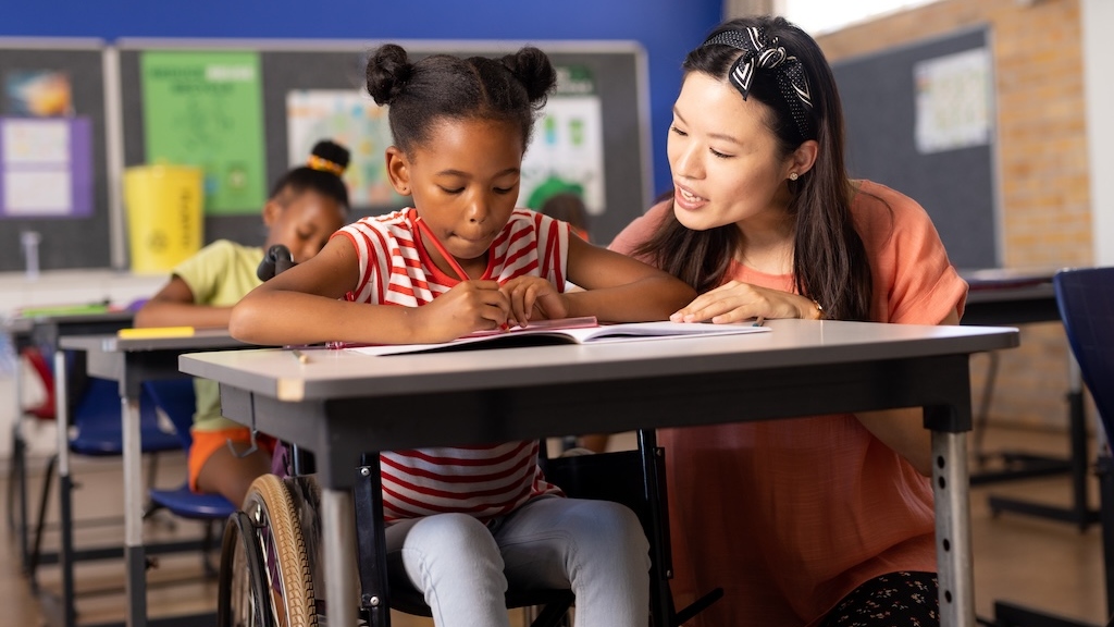 Photo of young girl sitting in school, getting help from her teacher in class. 