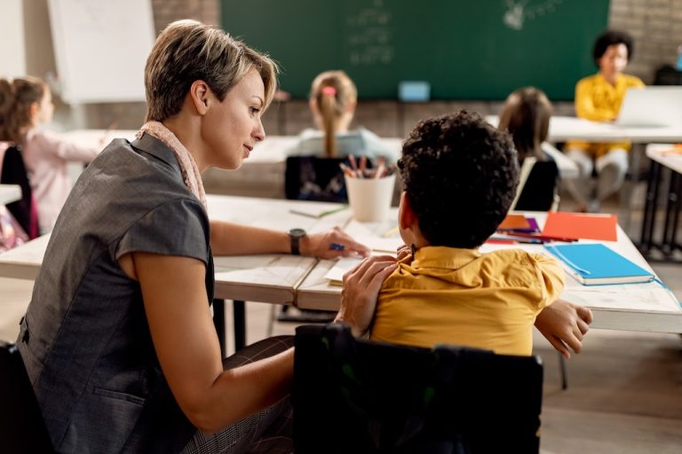 Female teacher talking to her elementary student while helping him with the assignment during a class at the school.