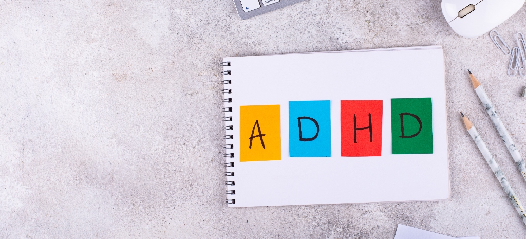 Letters "ADHD" written on a notepad laying on a desk, aerial view with pencils and computer mouse. 