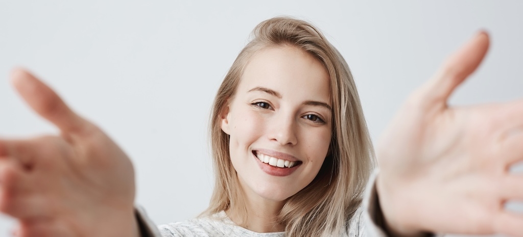 Headshot of pleasantlooking young Caucasian woman with broad smile stretching out arms to the camera. 