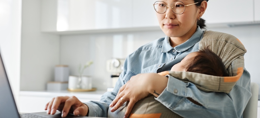 Young mom working on laptop while sitting with her baby in sling in the kitchen.