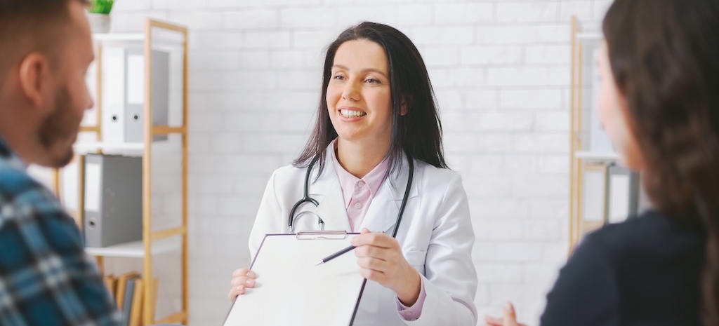 Female provider at neuropsychological testing facility is sitting with couple, holding a clipboard and pointing at it with a pen, discussing finances and cost of psych testing