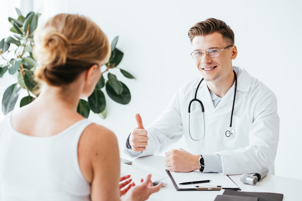 selective focus of cheerful doctor in glasses showing thumb up while having consult with patient