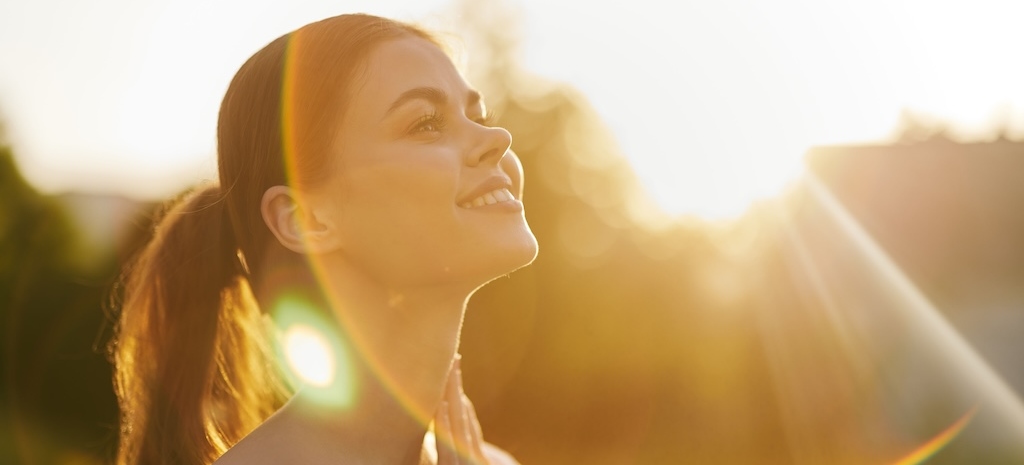 A young woman in a white off shoulder dress stands in nature, basking in the warm sunlight