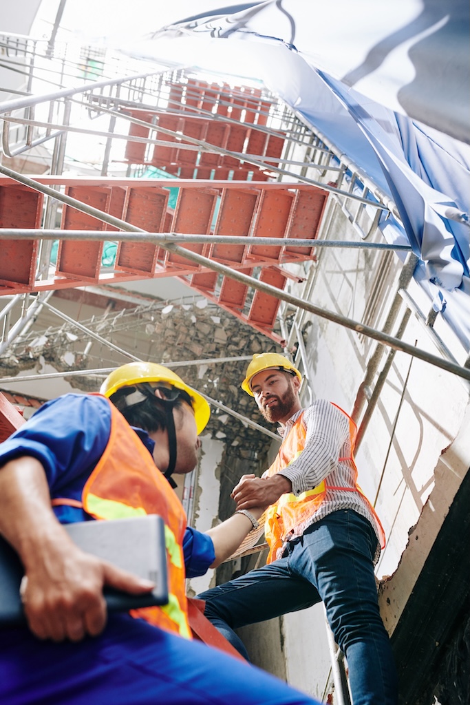 Builder in vest and hardhat helping coworker to climb up the scaffolding outside the building under construction
