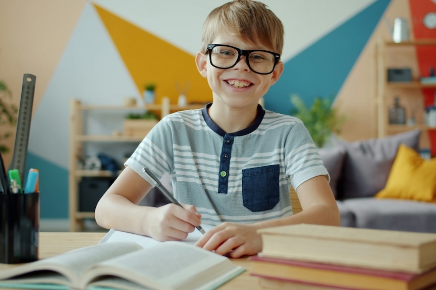 A young boy with glasses smiling while writing at his desk, representing children who may benefit from dysgraphia testing in San Diego.