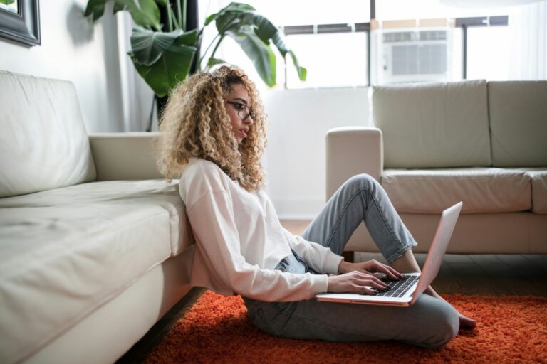 Young adult working on a laptop while sitting on the floor in a bright living room, appearing focused and thoughtful.
